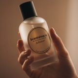 A hand holding a frosted glass bottle of Scentshop.io Achiever Essence Memory Scent with a black cap against a warm, solid brown background.