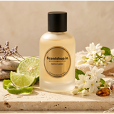 A frosted glass bottle of Scentshop.io Almond Blossom diffuser oil with a black cap, styled on a stone slab with lime slices, white blossoms, and a linen cloth.