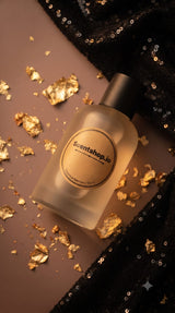 A frosted glass bottle with a black cap and a circular gold label reading 'Scentshop.io', styled on a warm brown surface with scattered gold leaf flakes and black sequined fabric.
